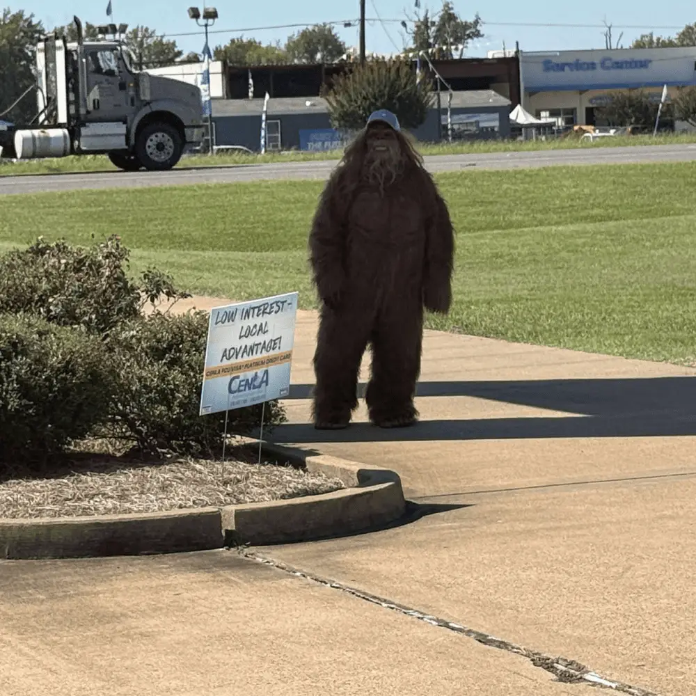 Bigfoot mascot in a brown costume standing on a sidewalk near a sign advertising low-interest local advantages, with a truck and buildings in the background, representing the playful branding of Bigfoot Electric & AC in Central Louisiana.