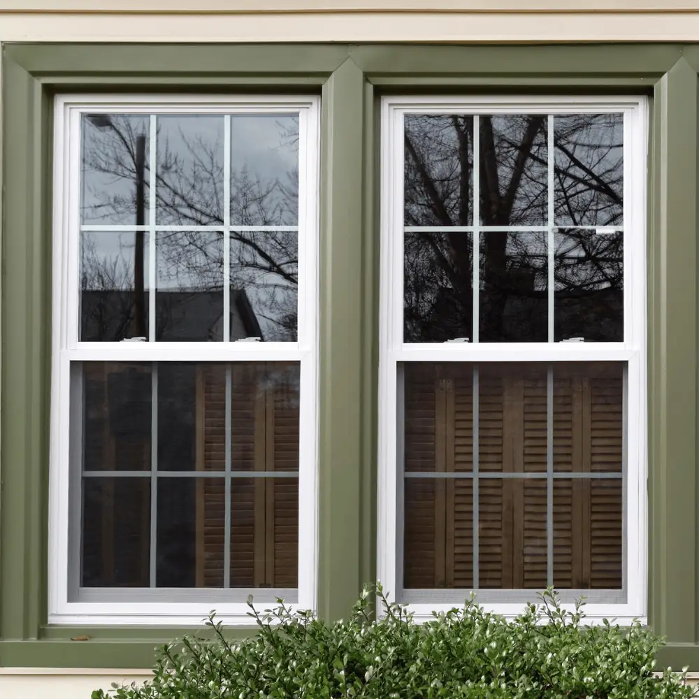 Replacement vinyl windows with green framing, showcasing reflections of trees and a nearby structure, emphasizing home improvement services by Bigfoot Electric & AC in Central Louisiana.
