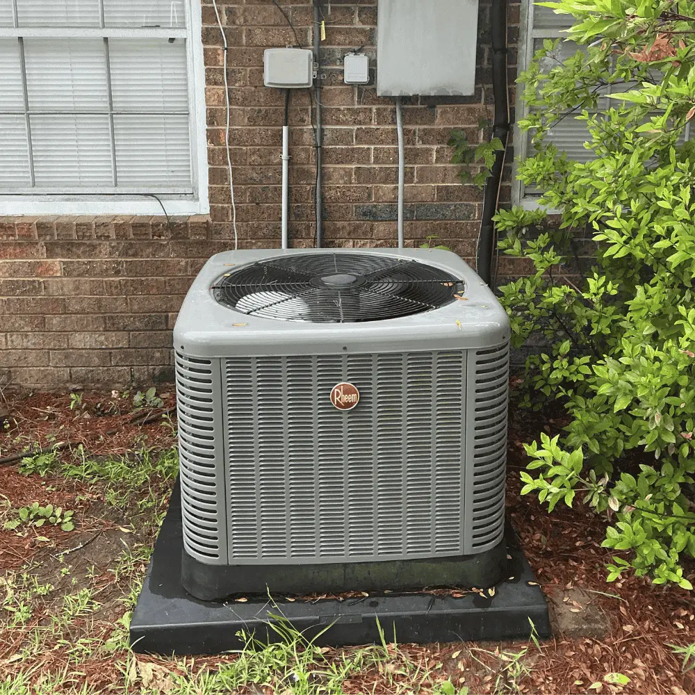 Rheem air conditioning unit installed outside a brick home in Central Louisiana, surrounded by greenery and utility connections, representing Bigfoot Electric & AC's HVAC services.