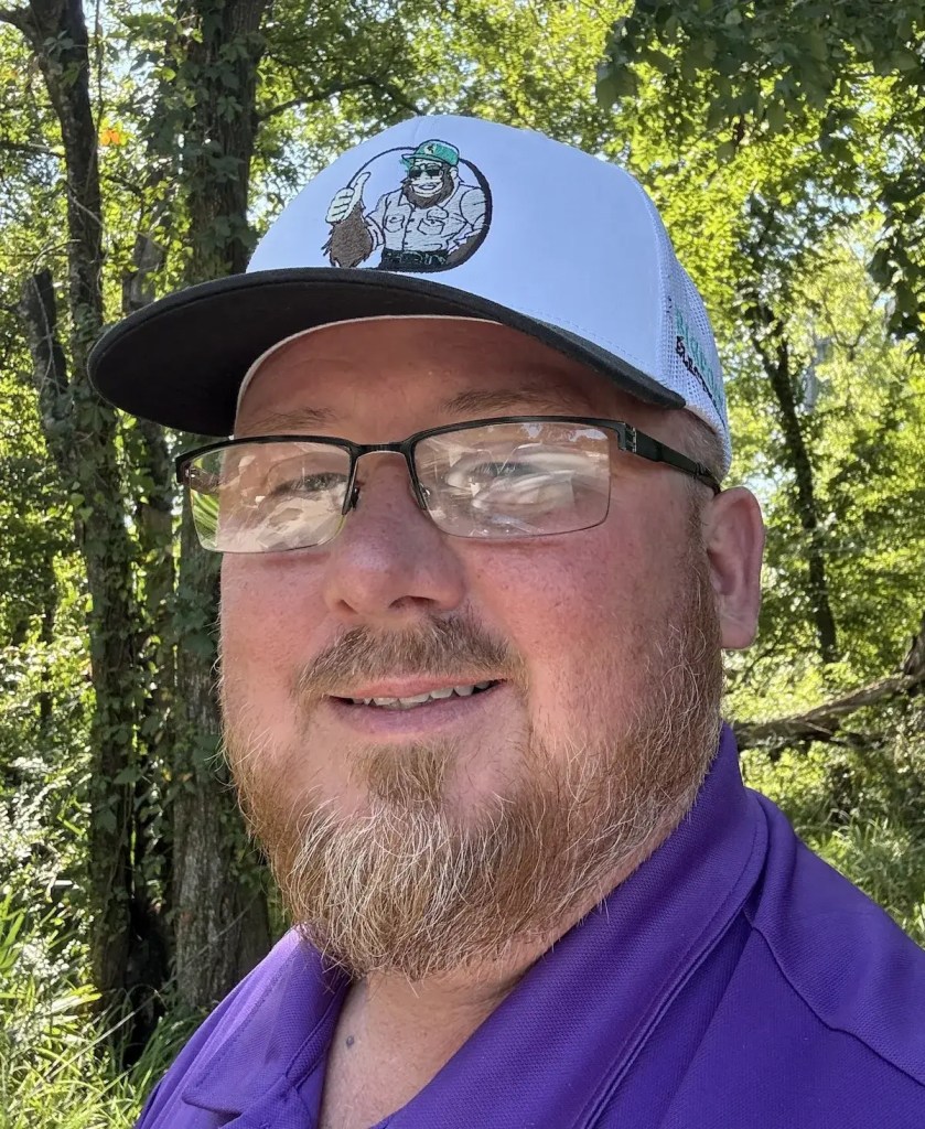 Man wearing a white cap featuring a cartoon Bigfoot logo, smiling outdoors with trees in the background, representing local service business Bigfoot Electric & AC.